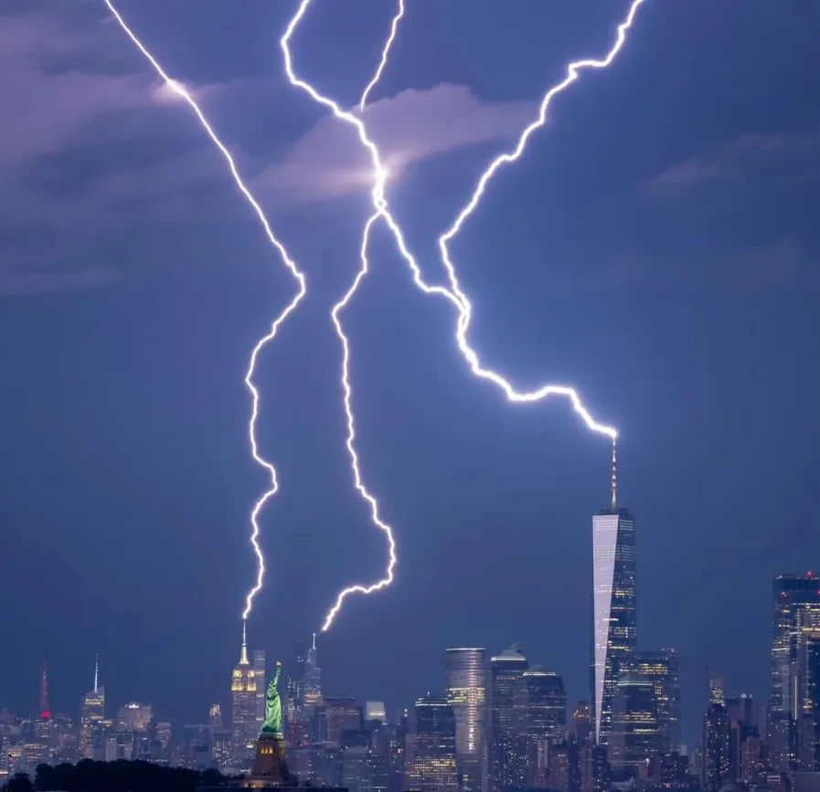 A dramatic nighttime photo of the New York City skyline with multiple bright lightning bolts striking down from the sky, one…