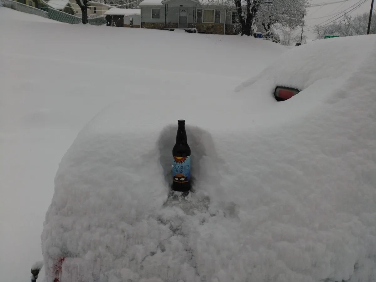 A dark beer bottle labeled 'Ninjabread Man' stands upright in a deep snowdrift, partially buried, with a snow-covered car…