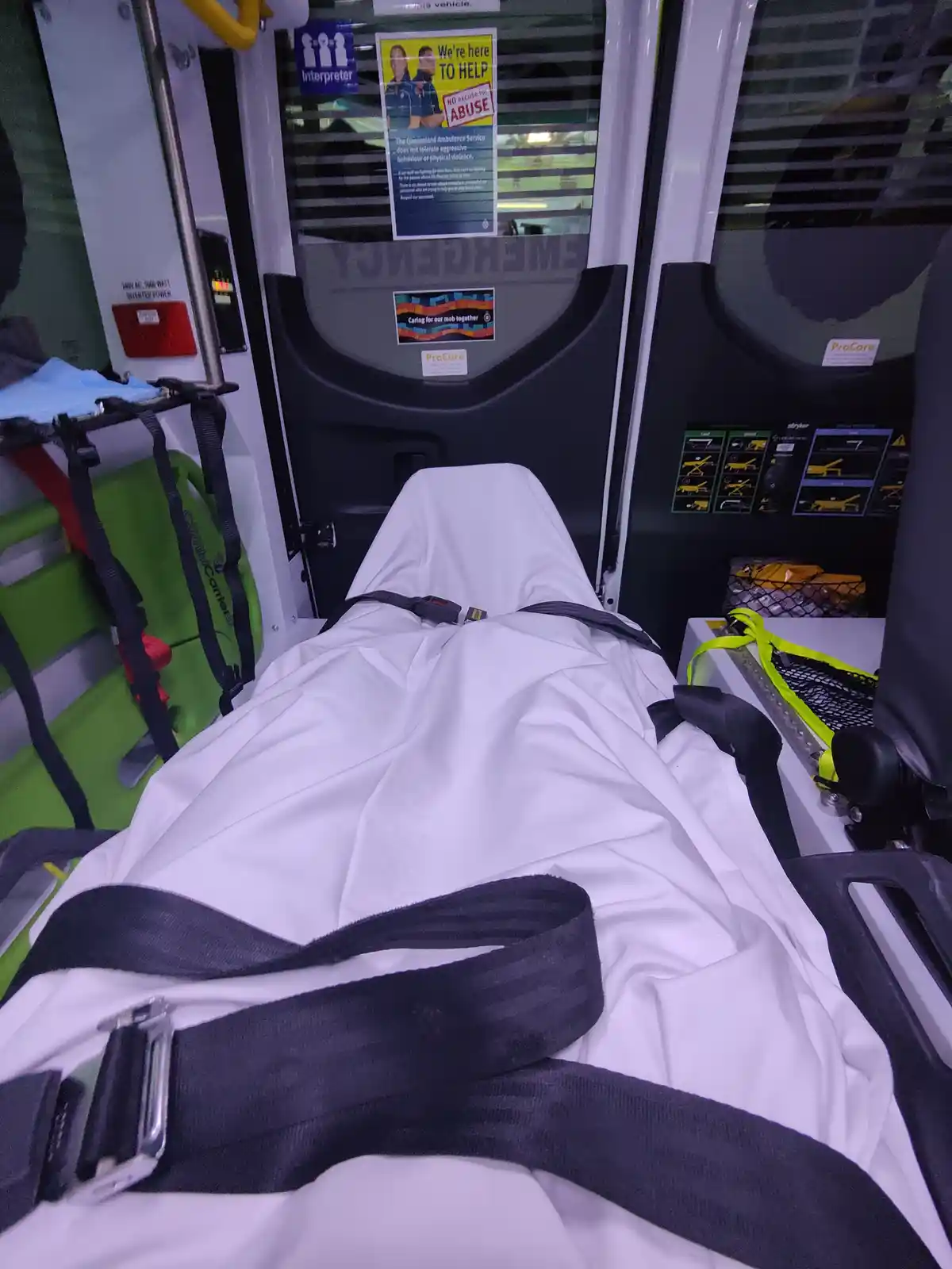 The image shows the interior of an ambulance with a patient stretcher covered in a white sheet and secured with black…