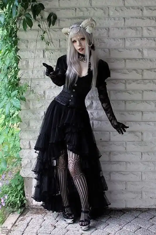 A person with long silver hair and small horns wears a black gothic outfit with lace gloves and striped stockings, posing…