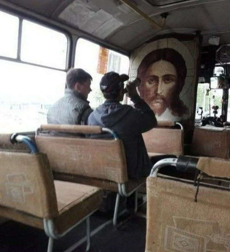 Inside a worn, public bus, two men sit facing forward; one appears to be taking a photo. Behind them, a large, faded…
