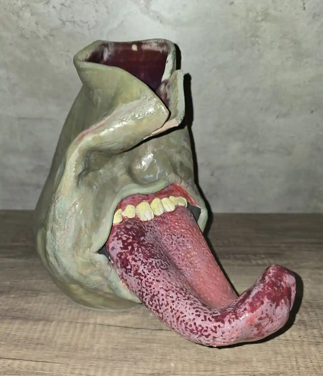 A ceramic vase shaped like a grotesque, open human mouth with visible teeth and a long, textured tongue sticking out…