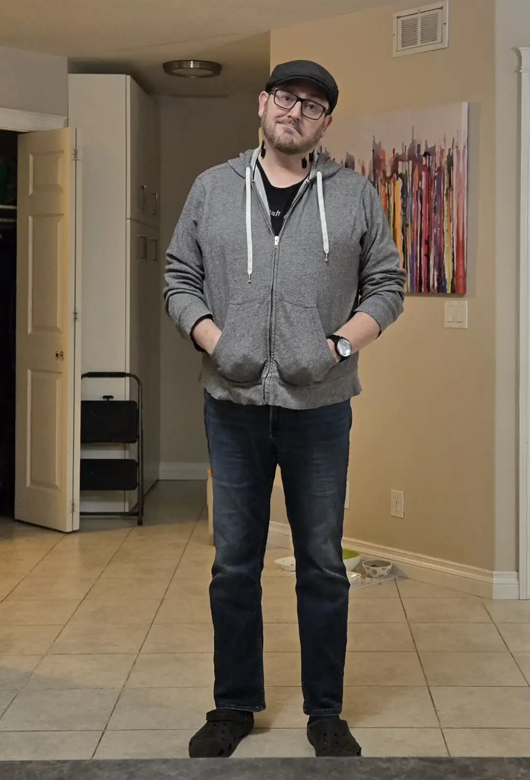 A man stands indoors wearing a grey zip-up hoodie, dark jeans, and black clogs, with his hands in his pockets and a smug…
