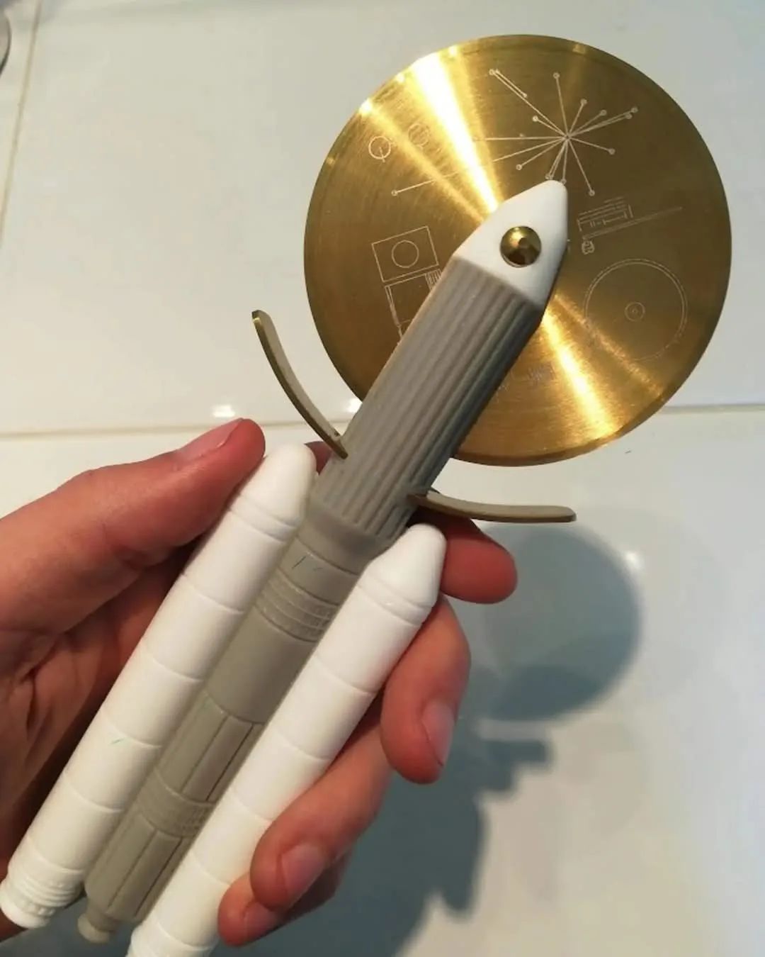 A hand holds a modified pizza cutter, with its circular golden blade serving as a rocket's nose cone and its handle forming…
