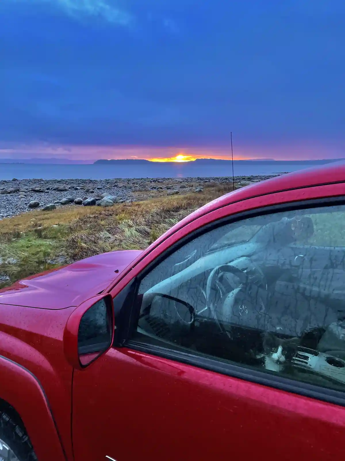 A red car is parked by a rocky shoreline at sunset, with raindrops on the window. The reflection in the wet glass shows a…