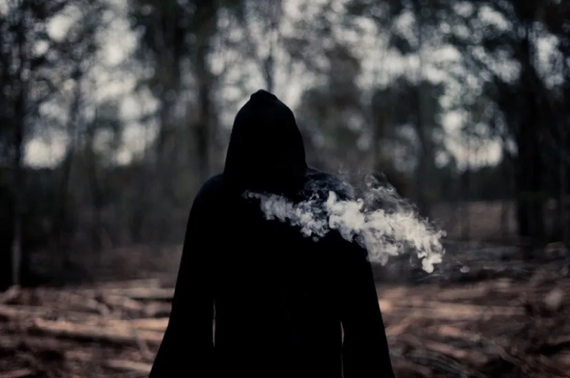 A person wearing a black hoodie stands with their back to the camera in a dim, wooded area. A large plume of white vapor or…
