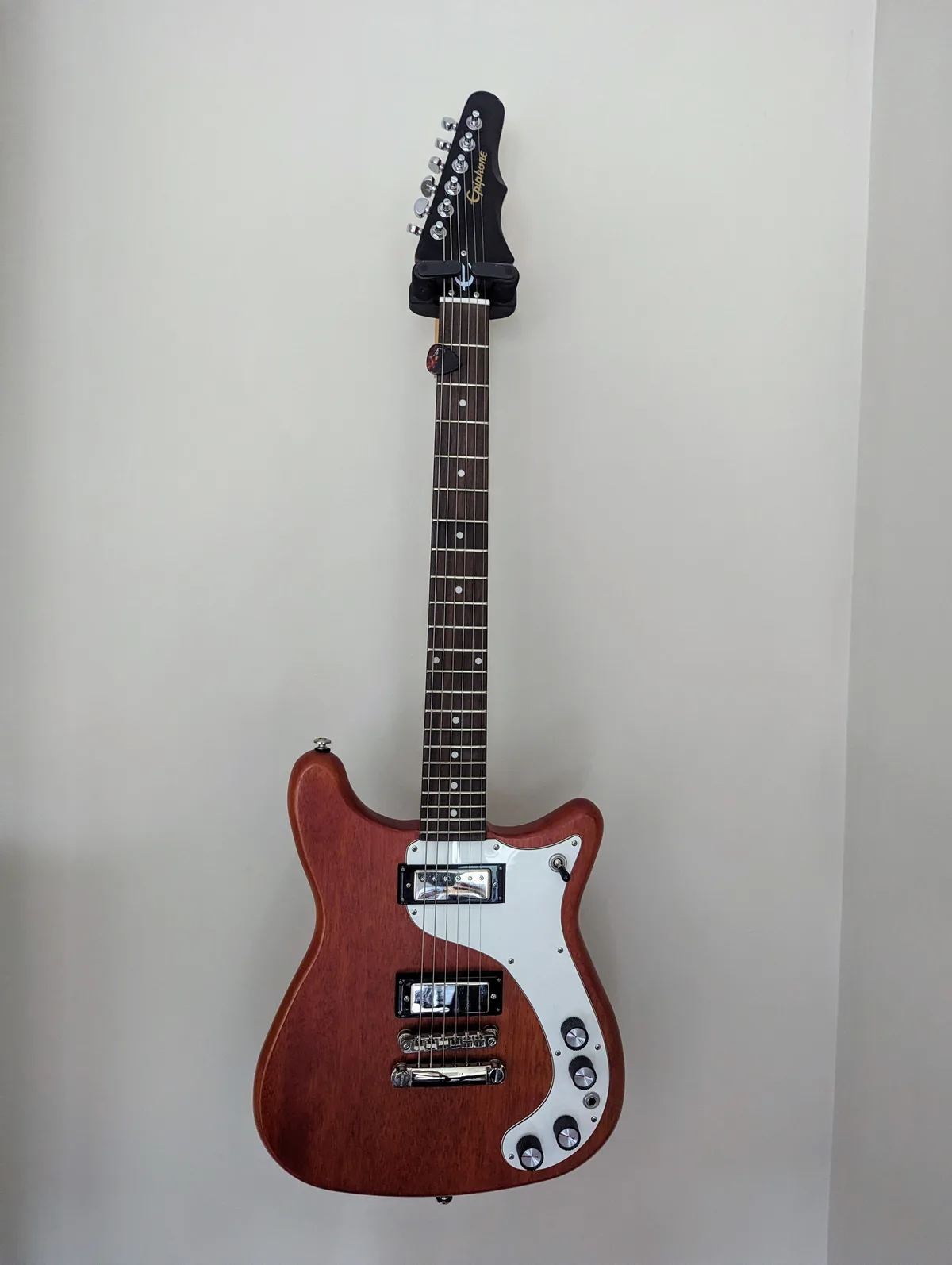 A brown electric guitar with a white pickguard and black hardware is mounted vertically on a plain white wall. The headstock…