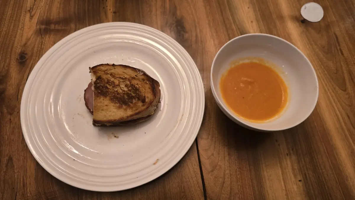 A partially eaten grilled sandwich on a white plate sits next to a bowl of orange soup on a wooden table, with the overlaid…