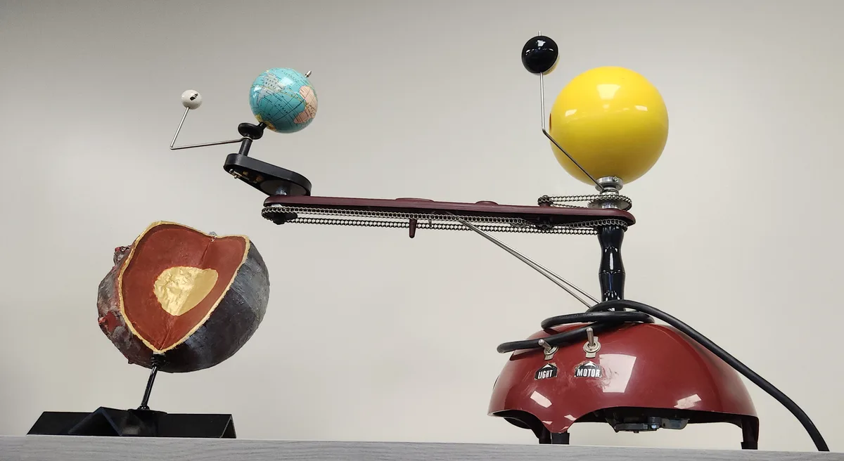 A mechanical model of the solar system with a yellow sun and blue Earth on a rotating arm, but the Earth is replaced with a…