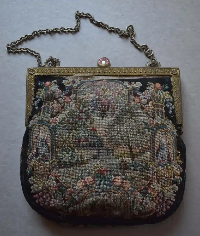 An ornate, vintage needlepoint purse with a gold frame and chain, featuring a detailed embroidered garden scene with two…