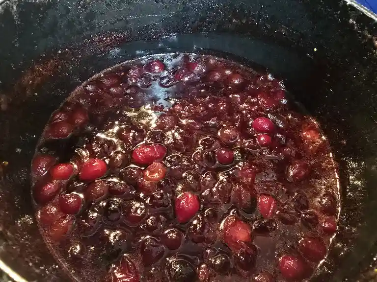 A close-up photo shows cranberries simmering in a dark sauce inside a black pot, with some berries bursting and others…