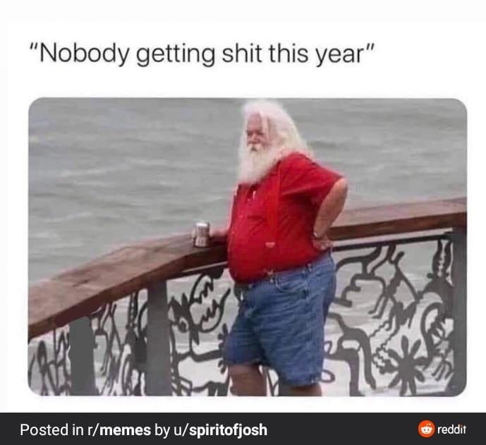 A man with a long white beard, wearing a red shirt and blue shorts, leans against a decorative railing overlooking water…