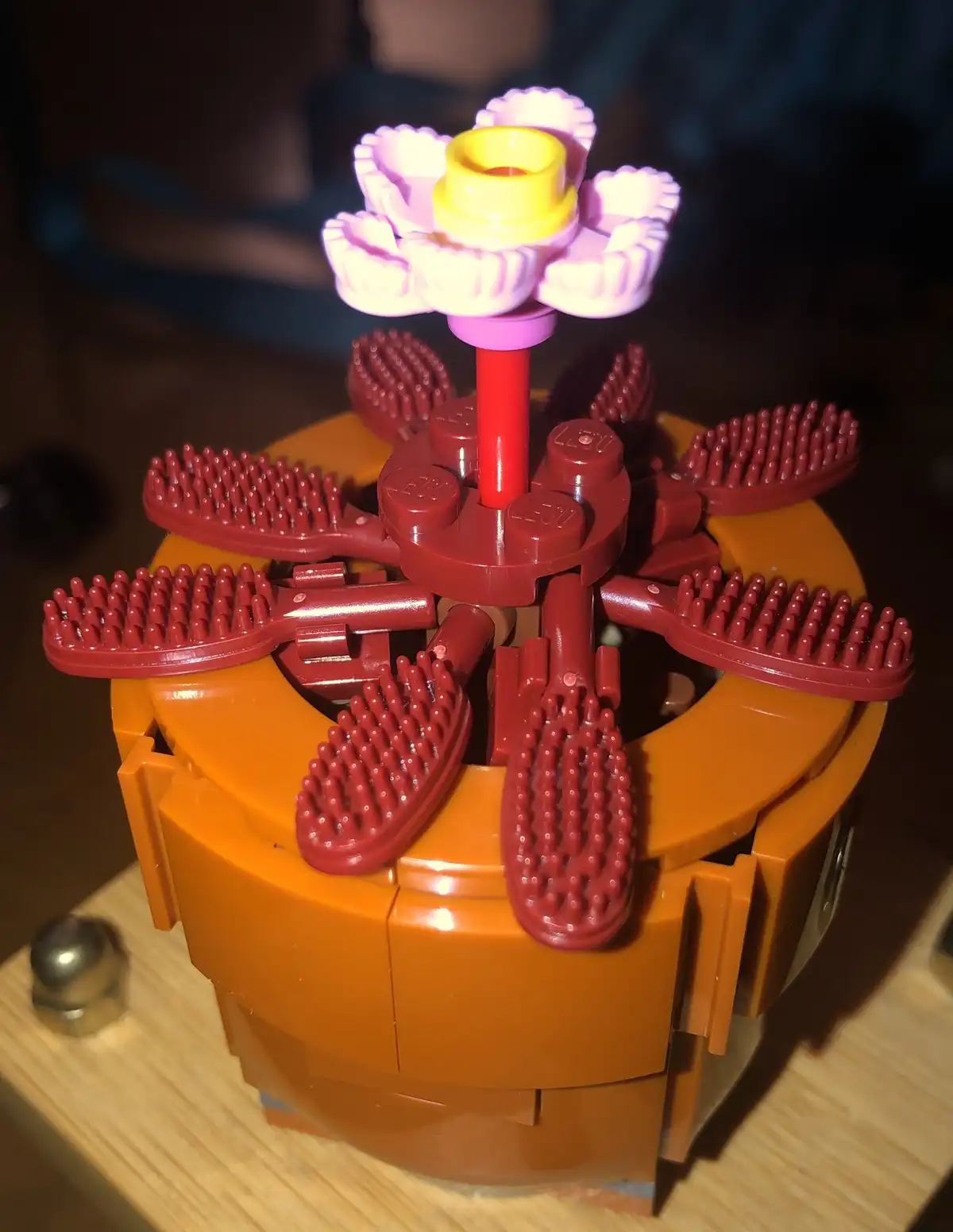 A close-up photo shows a LEGO creation resembling a flower, with a pink and yellow top, mounted on a red stem. Surrounding…