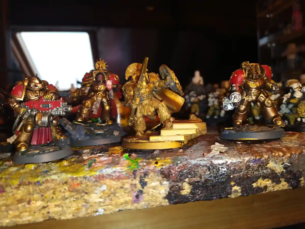 Several heavily painted, gold-accented fantasy or sci-fi miniatures stand on a paint-splattered wooden surface, with one…
