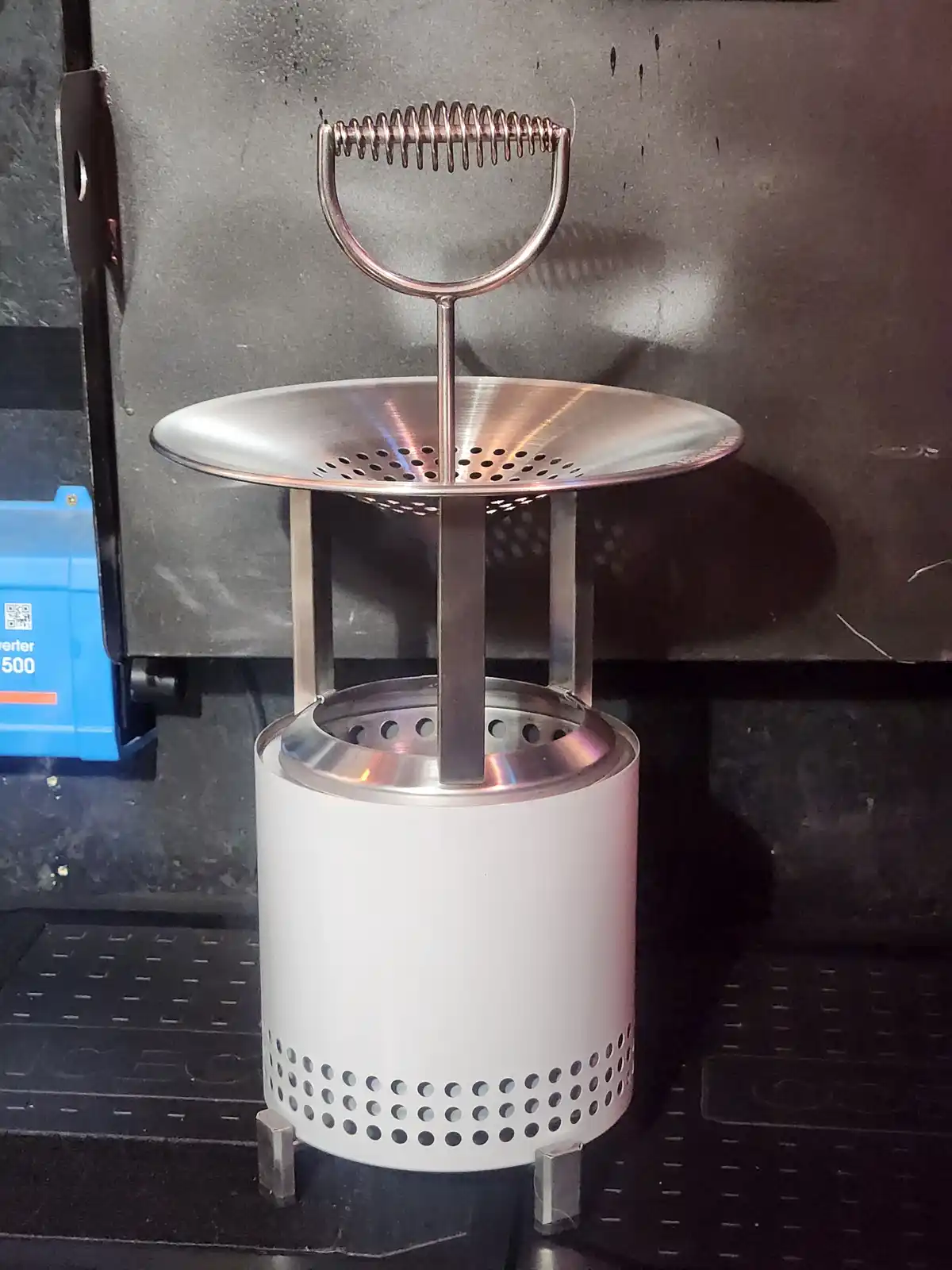 A metal strainer with a spring handle sits atop a white cylindrical base with perforations, placed on a black metal surface…