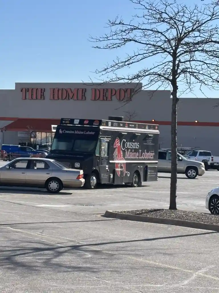 A black Cousins Maine Lobster food truck is parked in the lot of a Home Depot store on a sunny day. The truck features the…