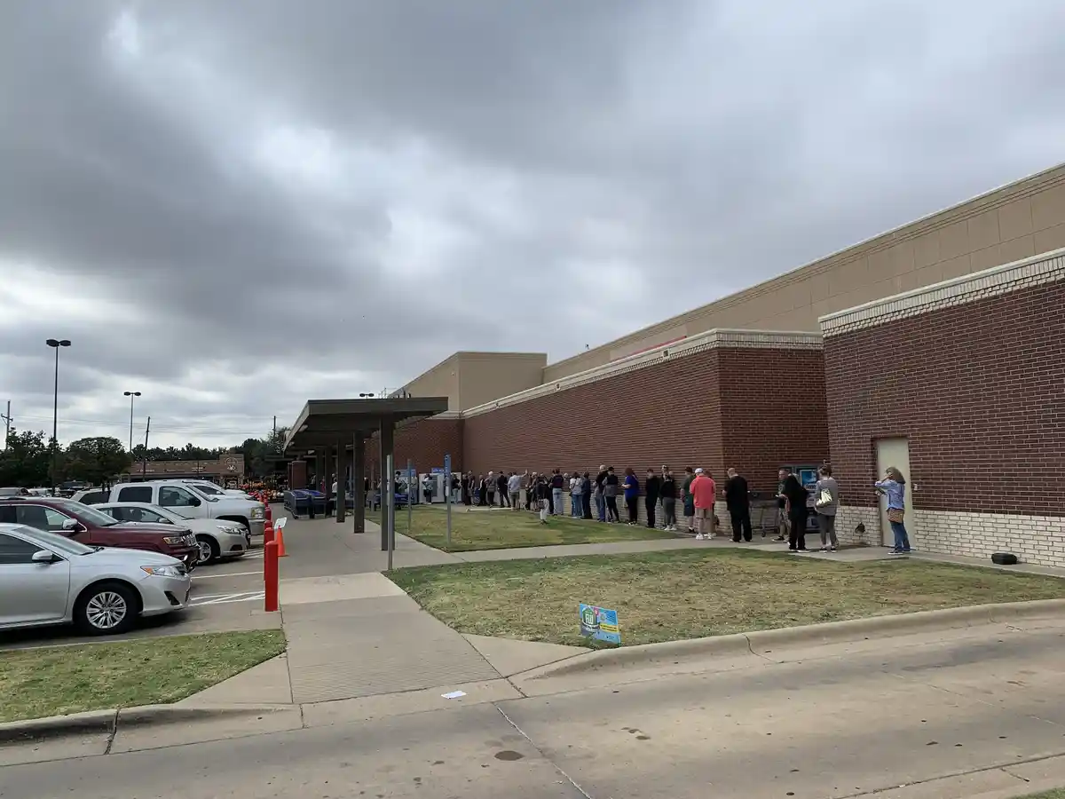 A long line of people stands outside a brick building under an overcast sky, with cars parked nearby. The overlaid text…
