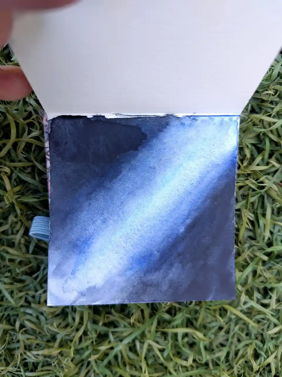 A hand holds open a small sketchbook or card, revealing a watercolor painting of a blue and black galaxy on the page. The…