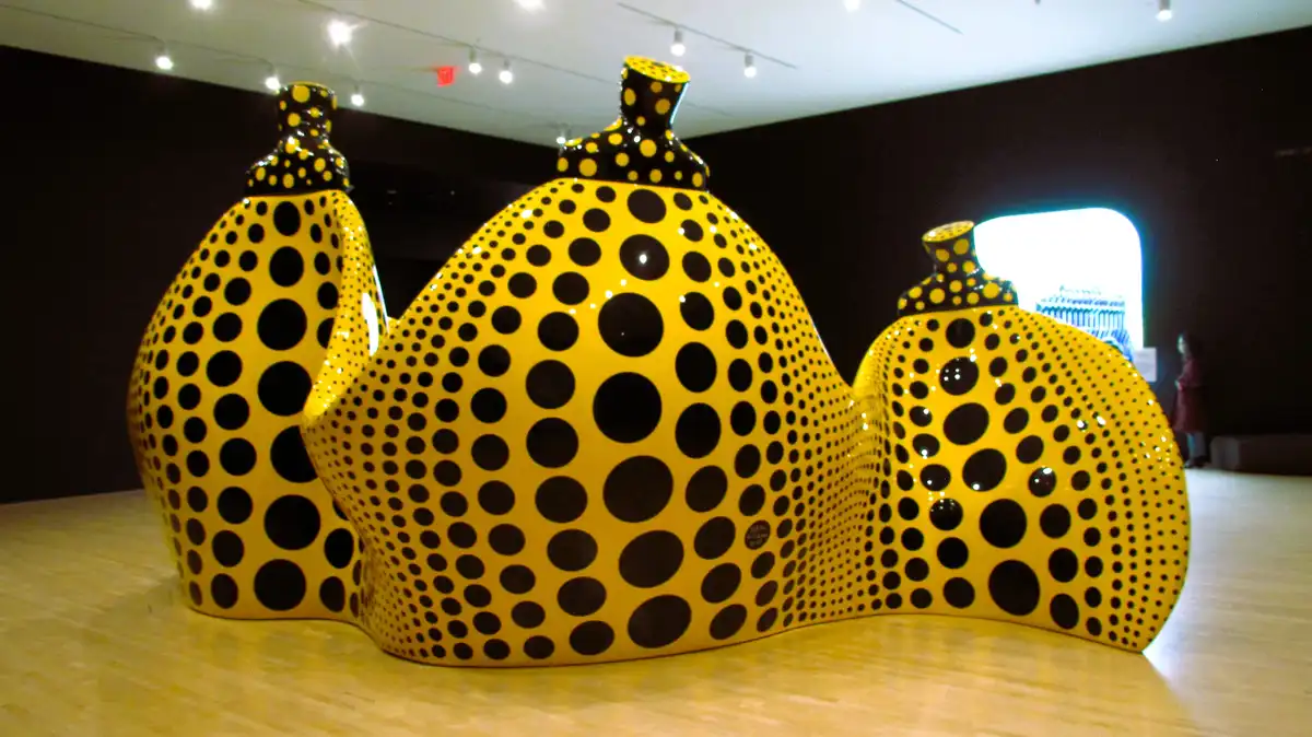 A large, yellow, wavy sculpture covered in black polka dots is displayed in a museum gallery. The sculpture has three…