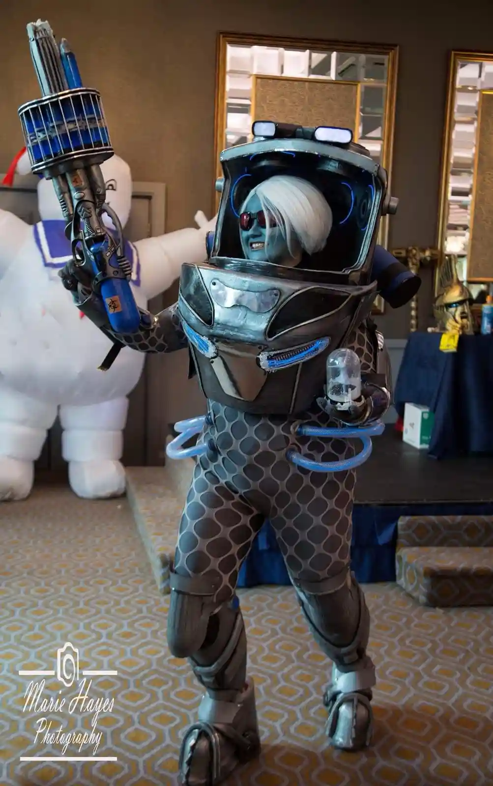 A person in a detailed, futuristic Ghostbusters cosplay with blue skin, white hair, and a large proton pack, stands in a…