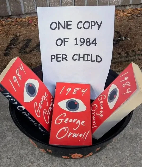 A black bucket with orange pumpkin designs sits outdoors on pavement, filled with three red copies of George Orwell's '1984'…