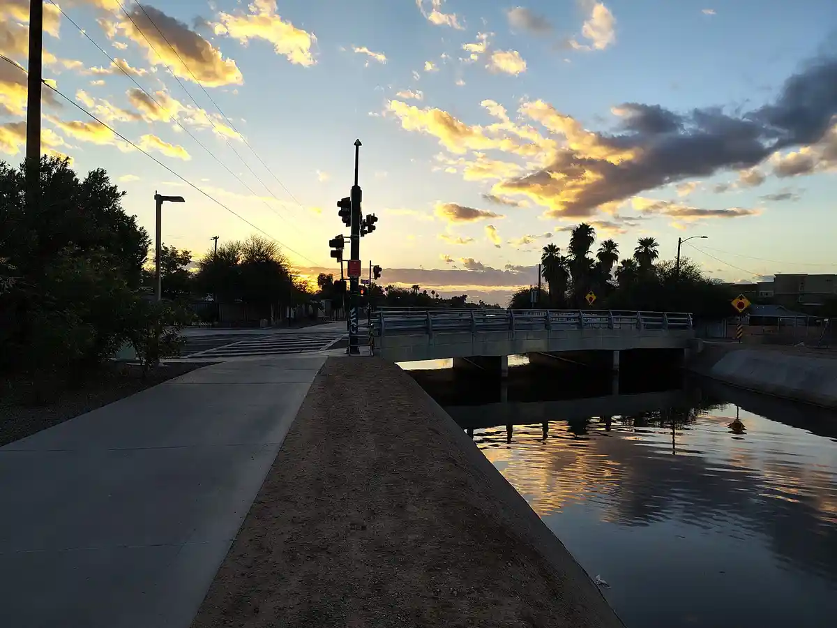 A serene sunset photo shows a concrete sidewalk running alongside a canal, with a bridge and traffic lights in the distance…