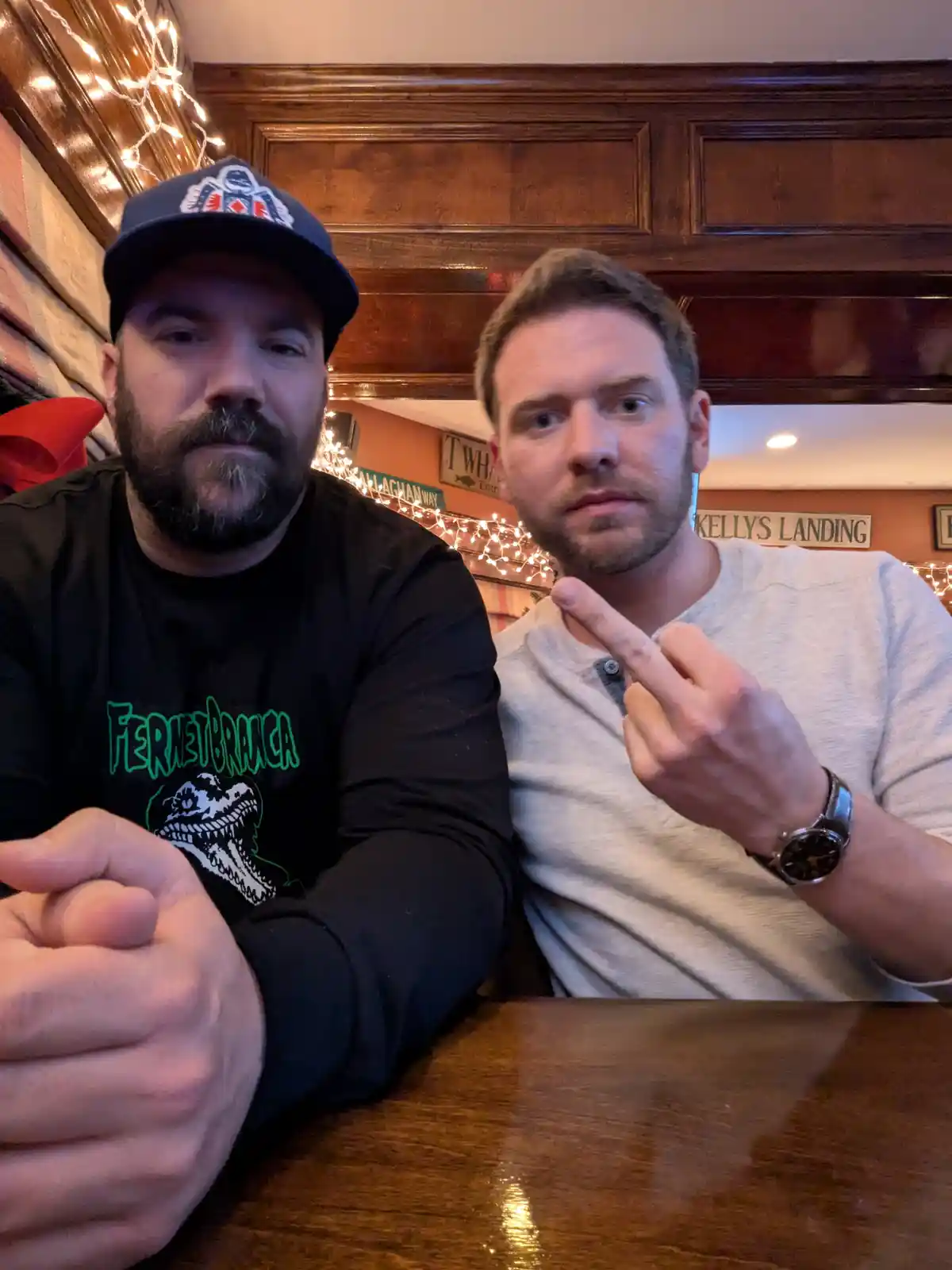 Two men sit at a wooden table in a warmly lit pub with string lights and wooden paneling. The man on the right, wearing a…