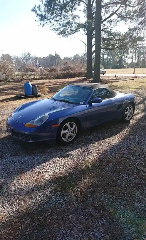 A blue Porsche Boxster convertible with a black soft top is parked on a gravel driveway surrounded by trees and dry grass…