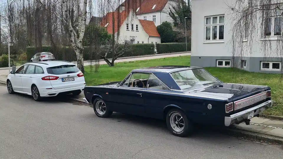 A dark blue classic American muscle car, a 1960s Plymouth Satellite, is parked next to a modern white Ford Mondeo station…
