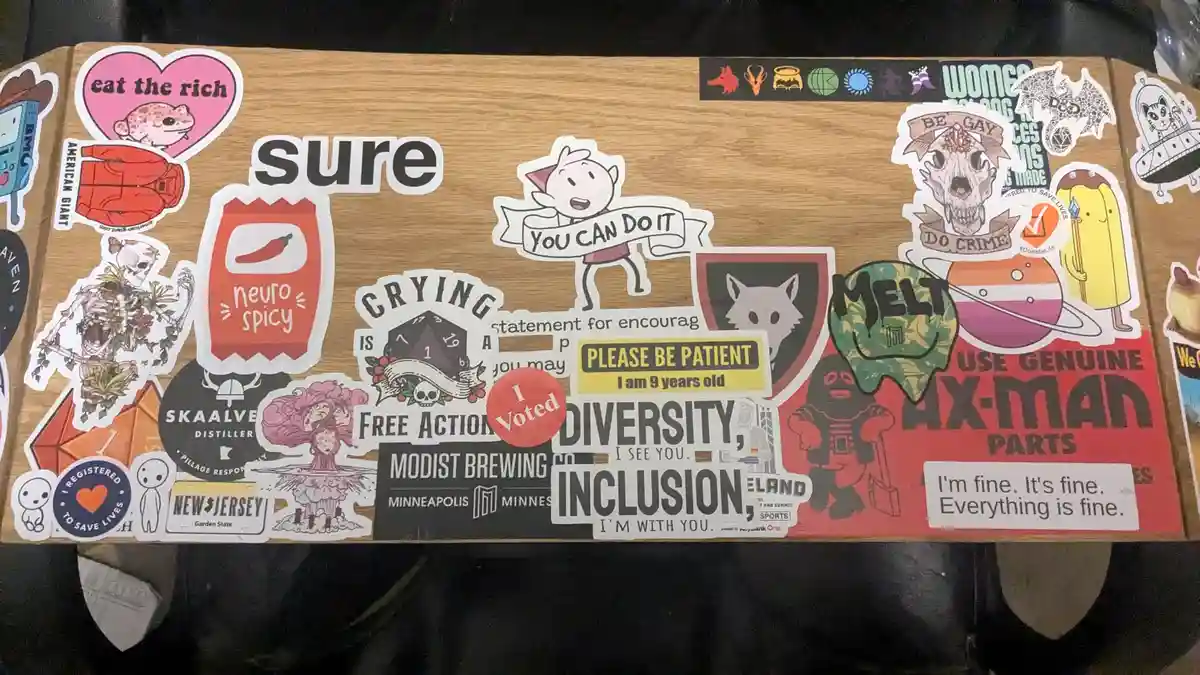 A wooden surface covered in numerous colorful stickers, including 'eat the rich', 'neuro spicy', 'I Voted', 'PLEASE BE…