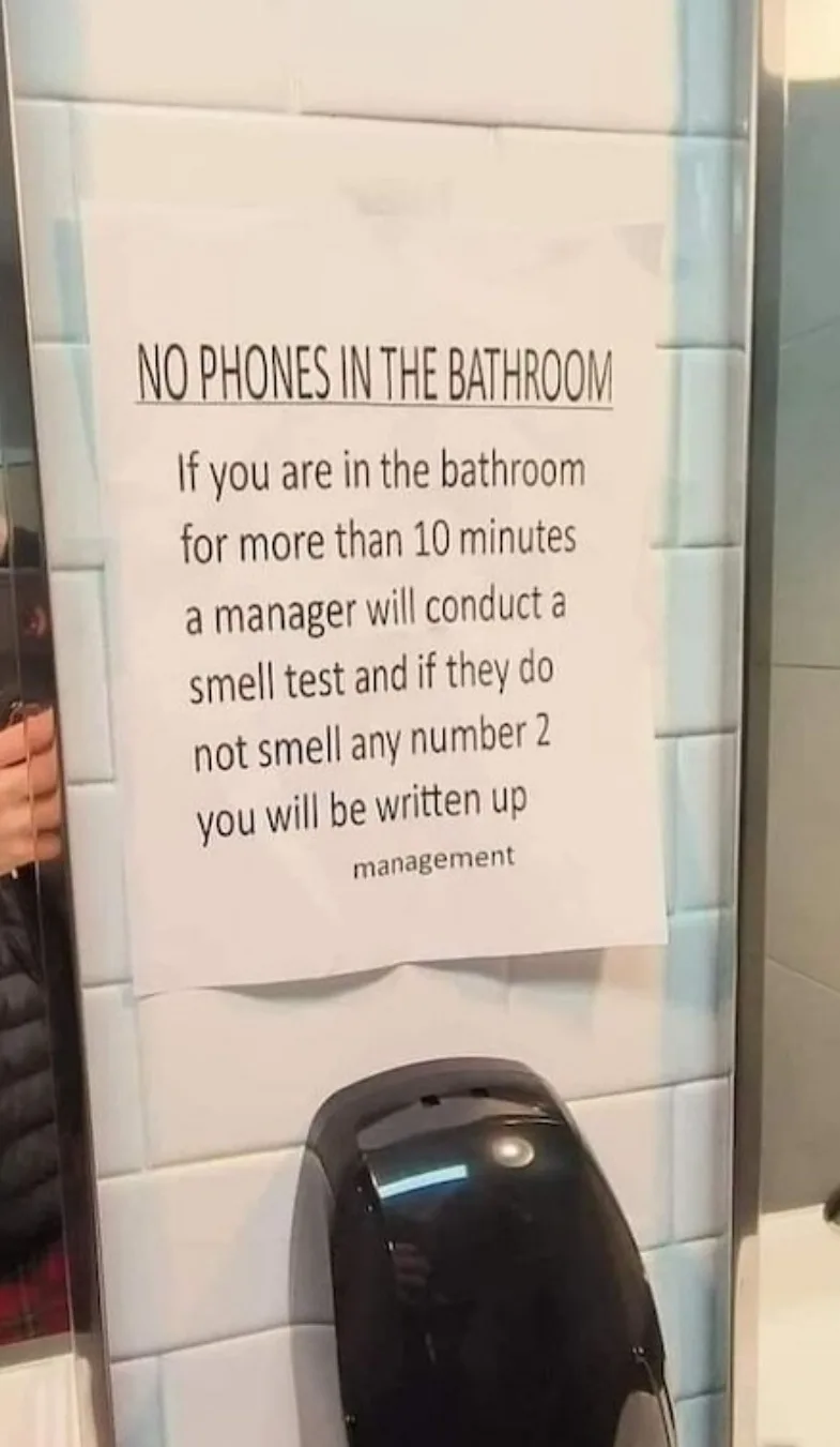 A white paper sign taped to a tiled bathroom wall next to a black hand dryer. The sign reads 'NO PHONES IN THE BATHROOM' and…