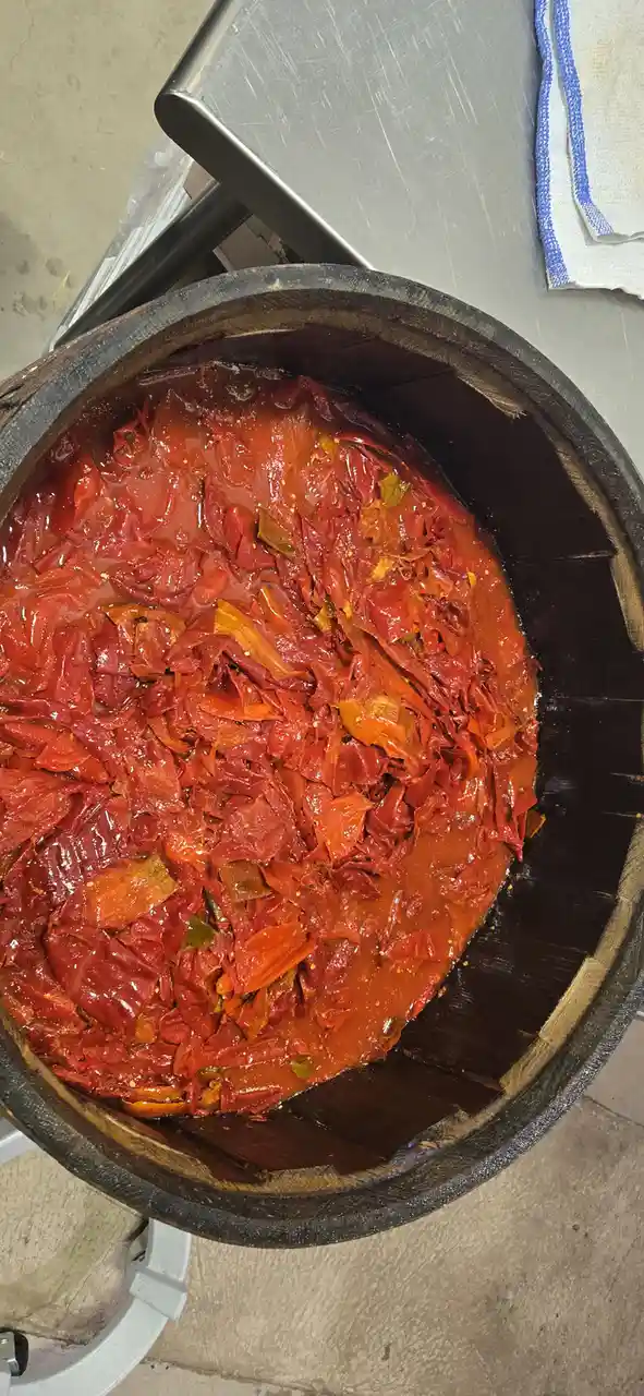A top-down view of a dark wooden barrel filled with a thick, red, pulpy mixture of what appears to be sun-dried tomatoes and…