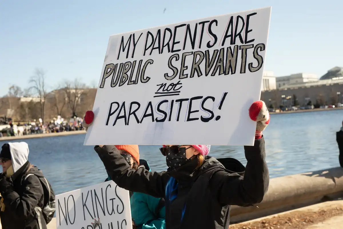 A person wearing a black jacket, pink beanie, and face mask holds a white protest sign with bold black and gray lettering…