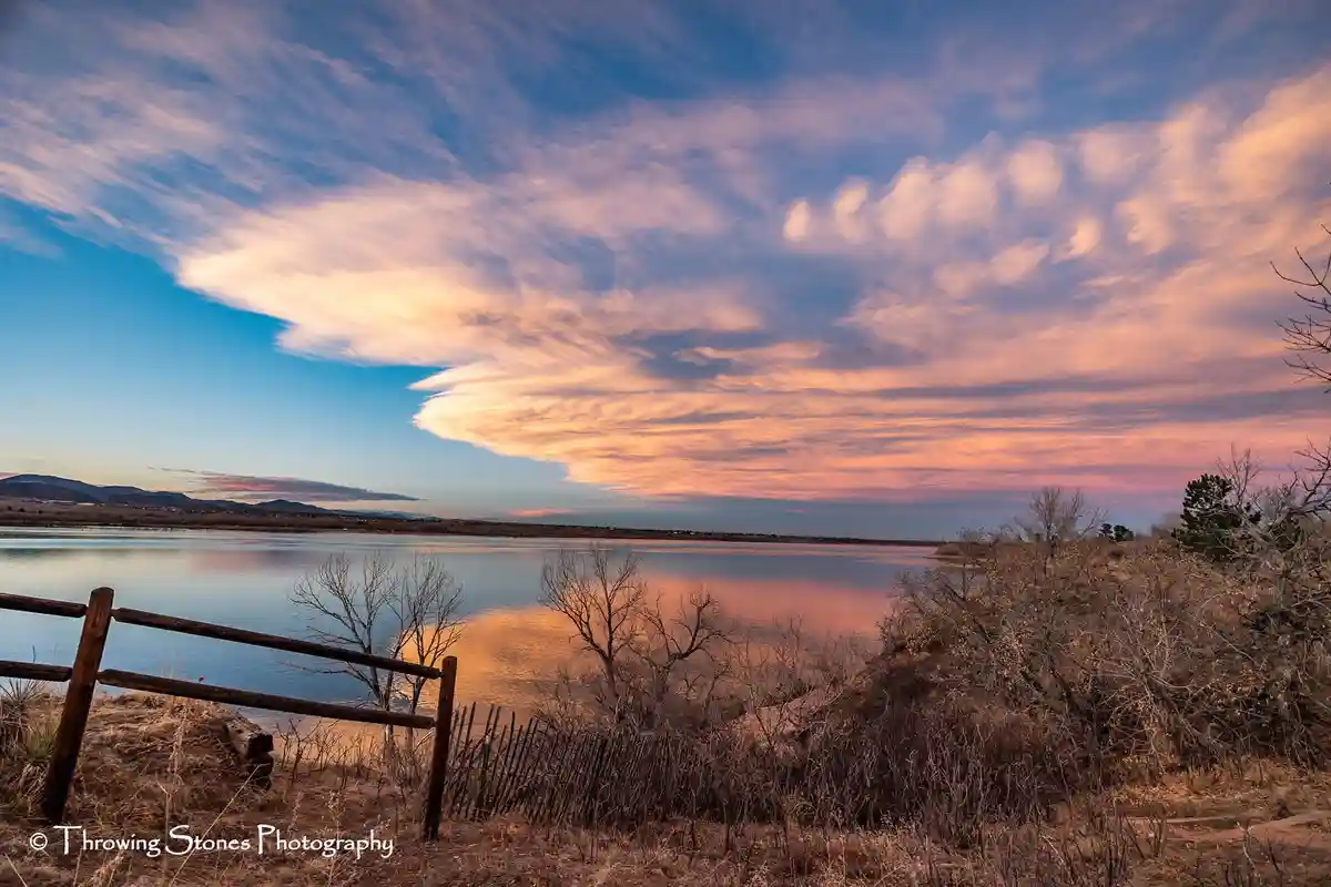 A serene landscape photo of a lake at sunset with vibrant orange and pink clouds reflected in the water, framed by bare…