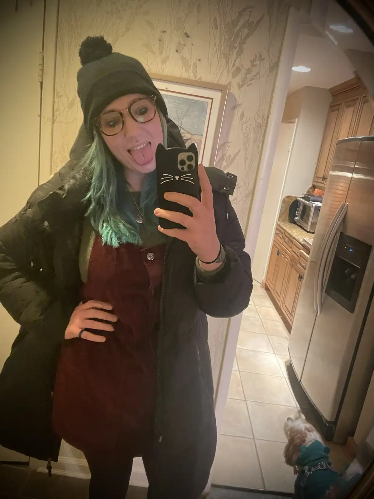 A person with green hair, glasses, and a black beanie takes a mirror selfie in a kitchen, sticking their tongue out while…