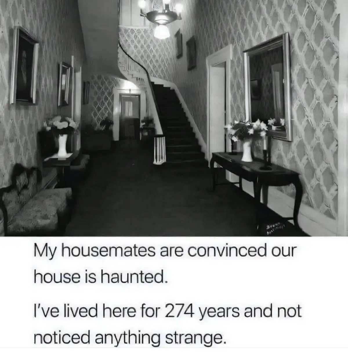 A black-and-white photo of an old, ornate hallway with patterned wallpaper, a staircase, framed pictures, and furniture…