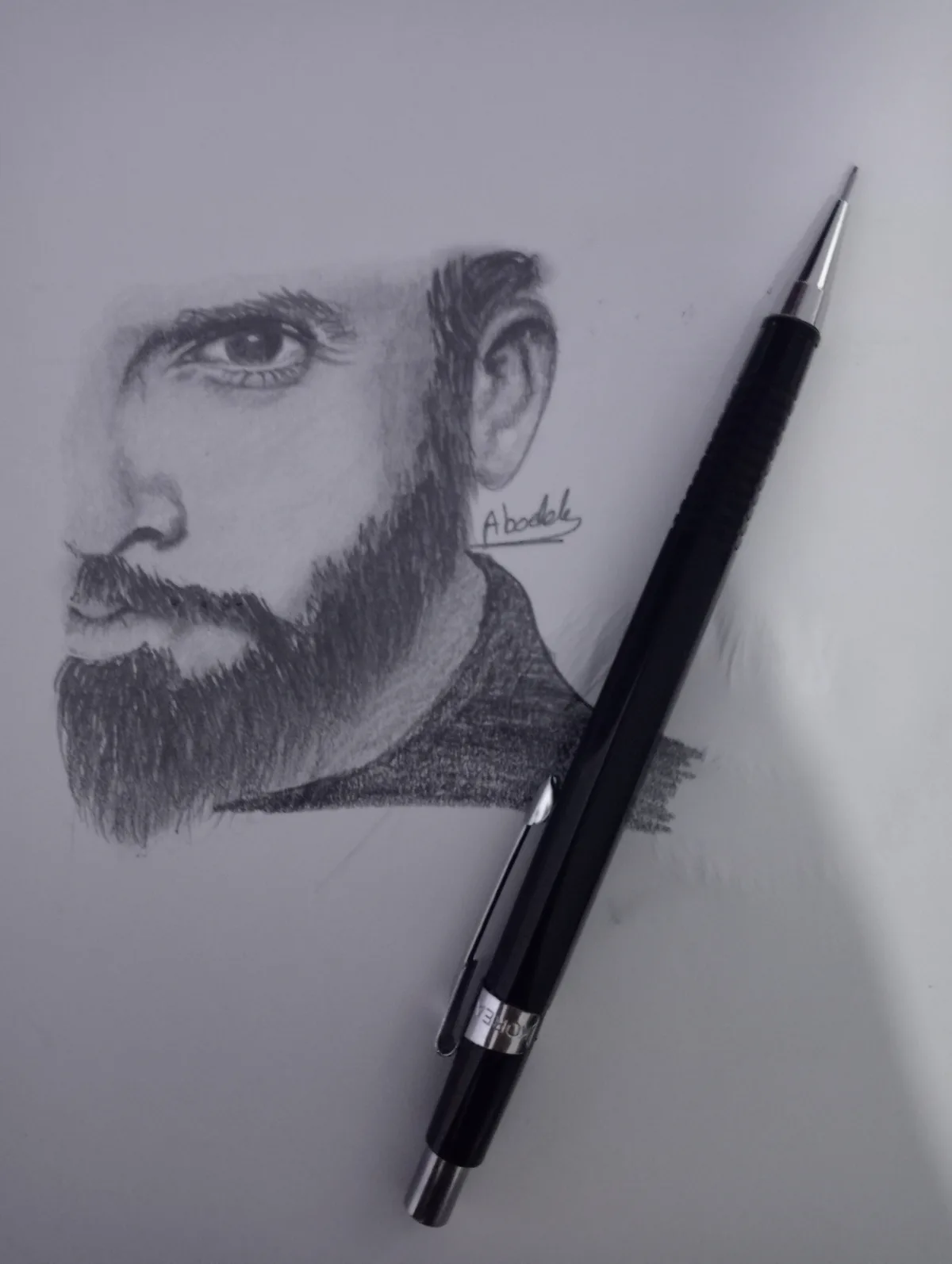 A detailed pencil sketch of a man's face with a beard and intense gaze, signed 'Abodele', lies next to a black mechanical…