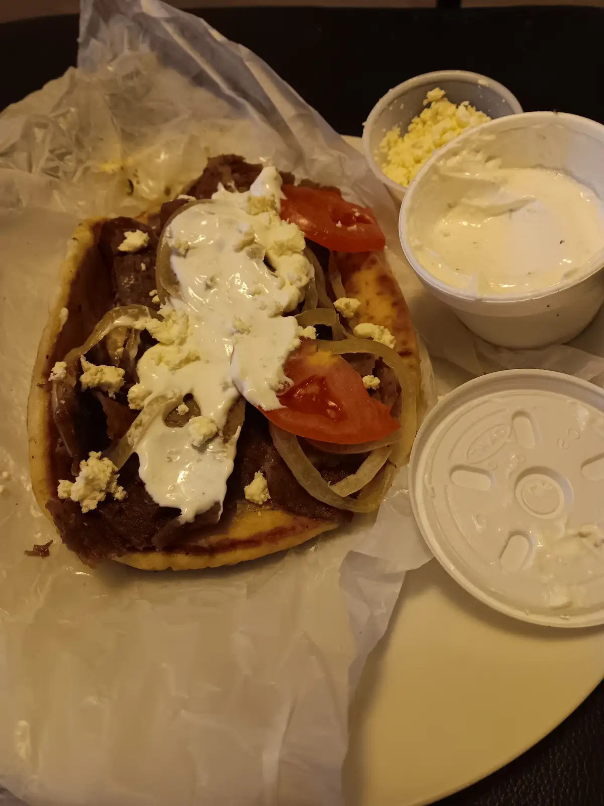 A close-up photo shows a gyro sandwich on a pita bread, topped with sliced meat, tomato, onion, crumbled feta cheese, and a…