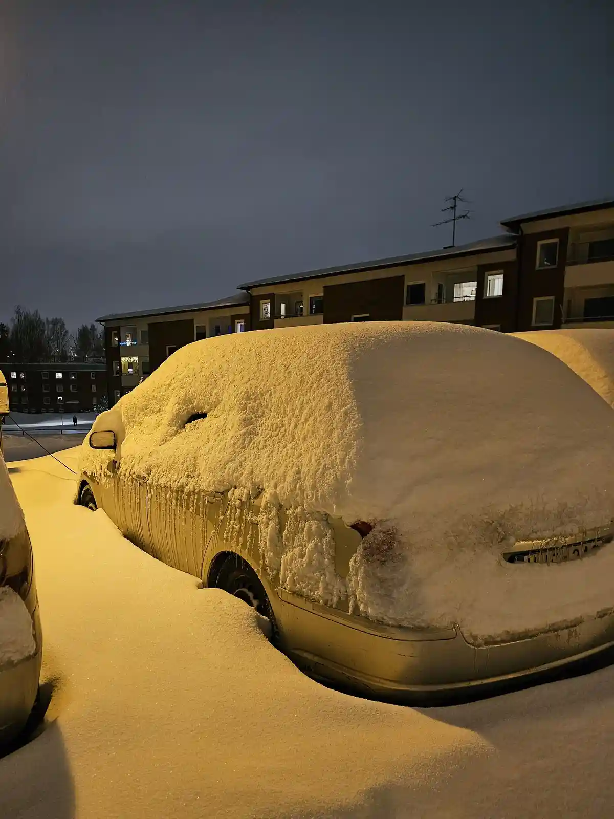 A car is almost entirely buried under a thick layer of snow in a parking lot at night, with only parts of the rear bumper…