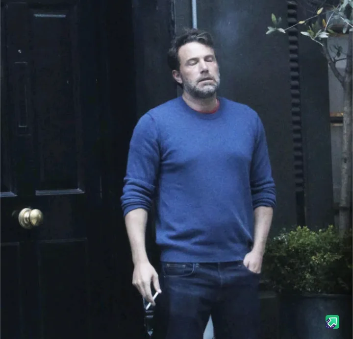 A man with a beard and short hair, wearing a blue sweater and dark jeans, stands outside a dark door with his eyes closed…