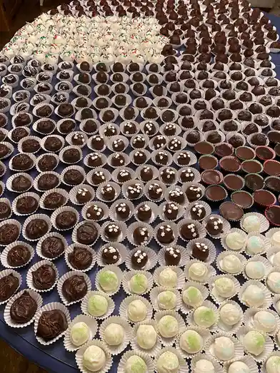 A large, round table is covered with hundreds of individually wrapped chocolate truffles arranged in neat rows. The truffles…