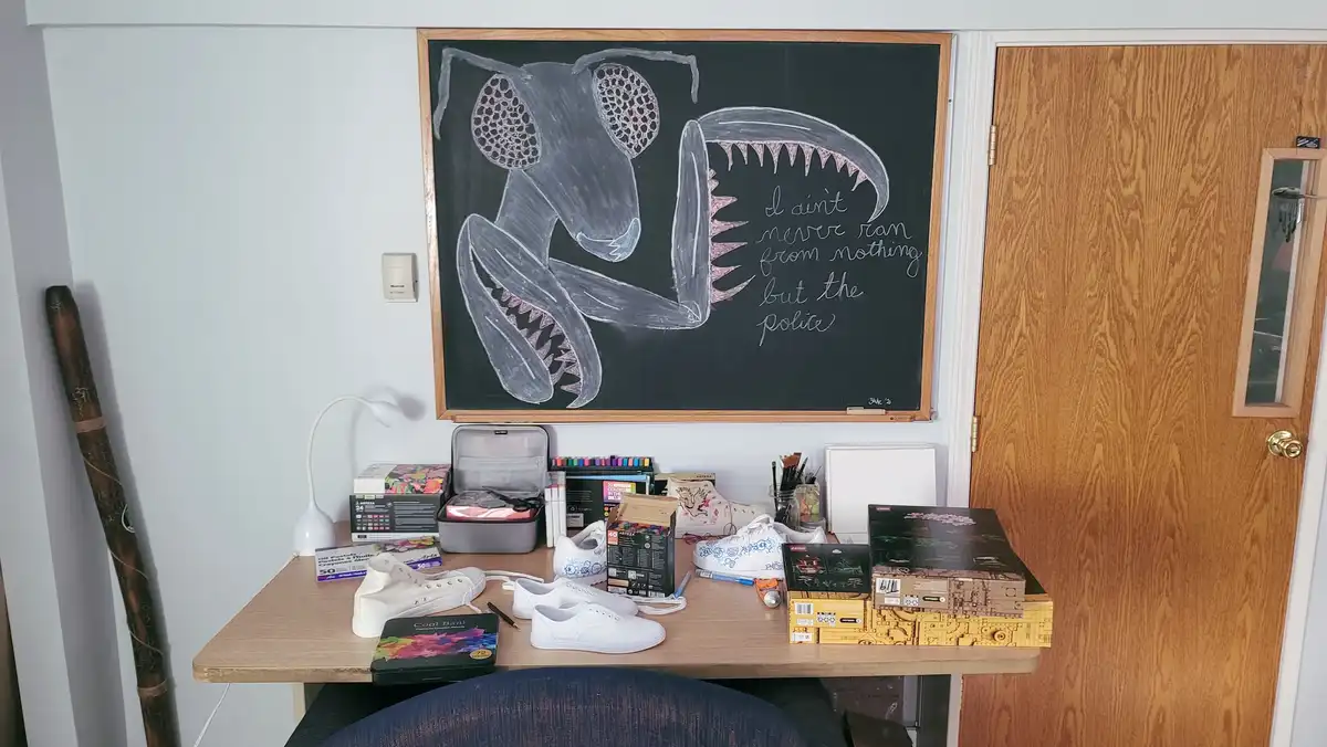 A chalk drawing of a monstrous creature with large eyes and sharp teeth is on a blackboard above a cluttered desk. The…