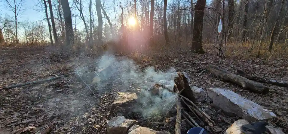 A campfire pit in a forest with smoke rising, surrounded by rocks and fallen leaves, with the sun setting behind bare trees…