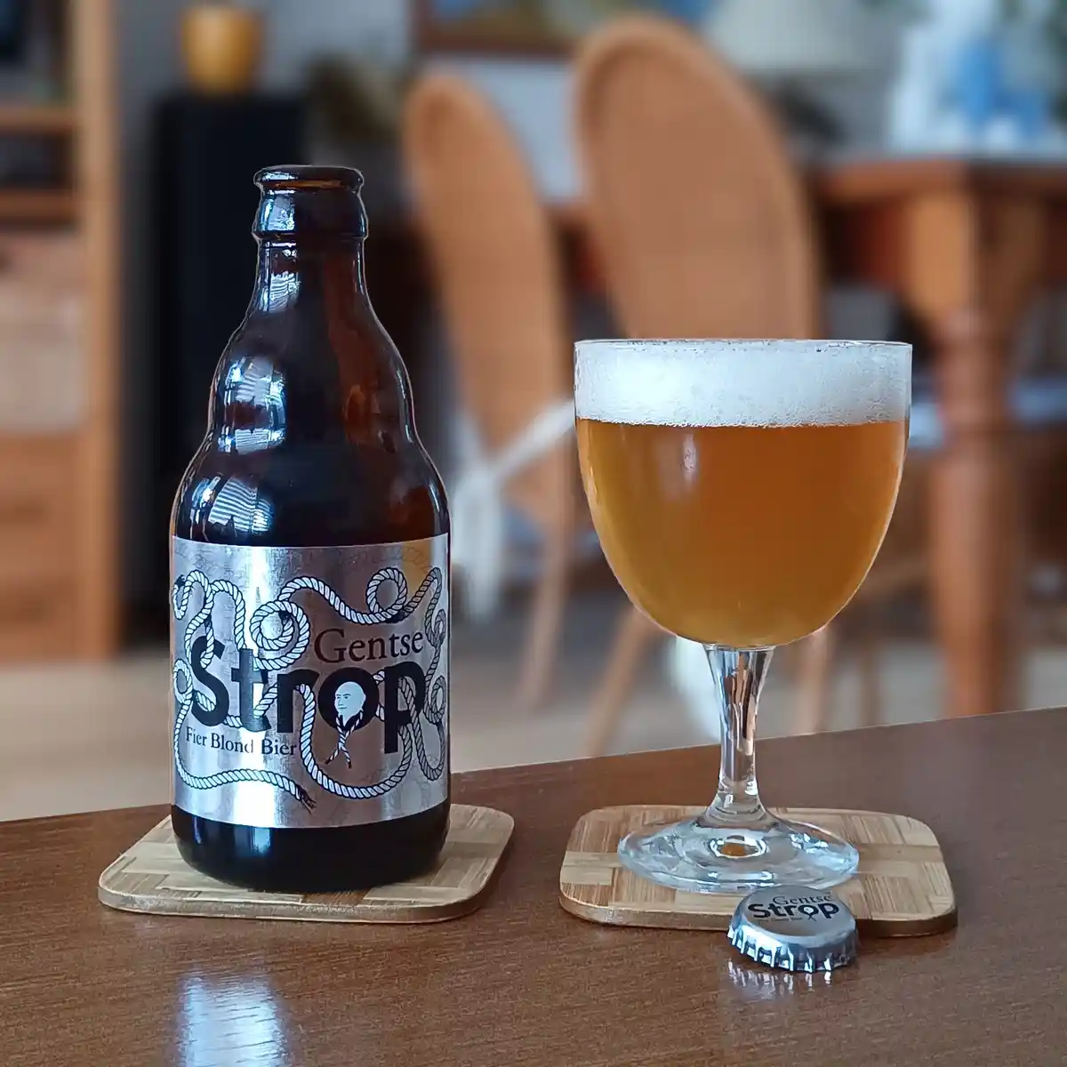 A brown beer bottle labeled 'Gentse Strop' with rope graphics sits next to a stemmed glass filled with golden beer and white…
