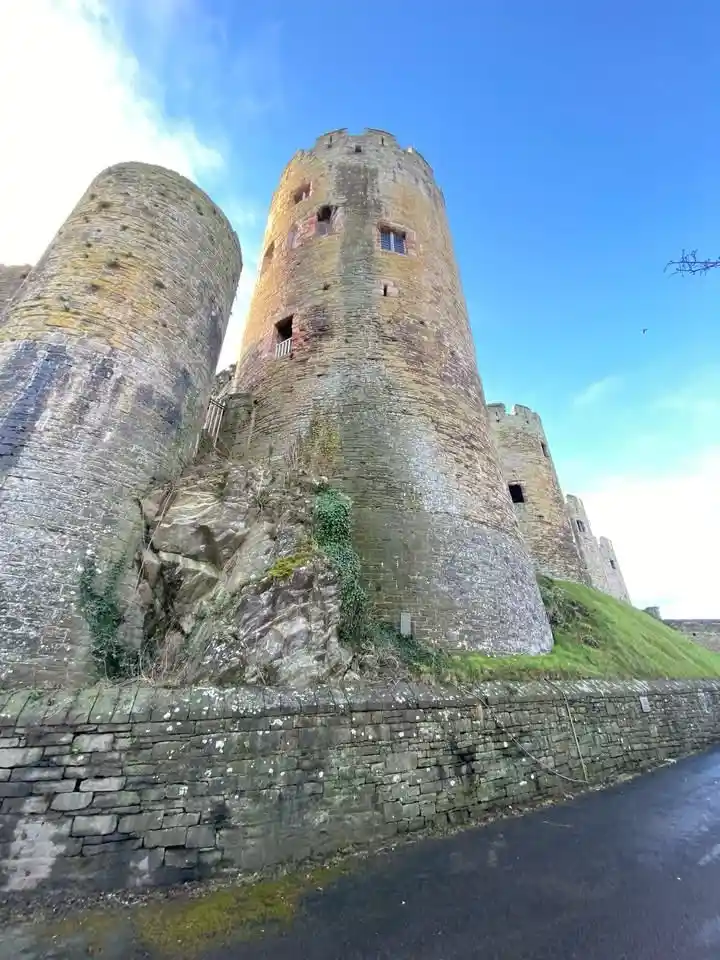 A low-angle photo shows massive, ancient stone castle towers rising against a bright blue sky, with ivy growing on the rocks…