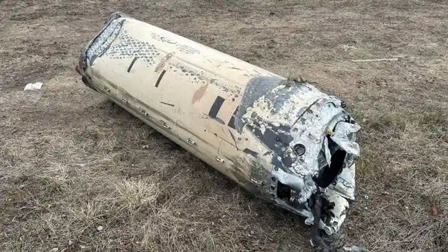 A damaged, cylindrical fuselage section of an aircraft lies on dry grass, showing burn marks, dents, and missing panels. The…