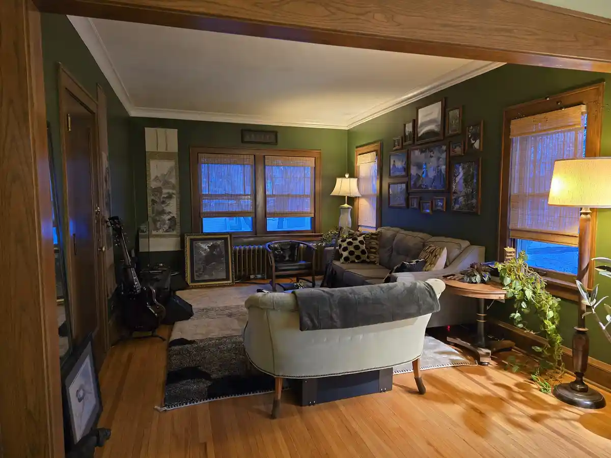 A cozy, well-decorated living room with green walls, wooden floors, and multiple framed pictures on the walls. A gray sofa…