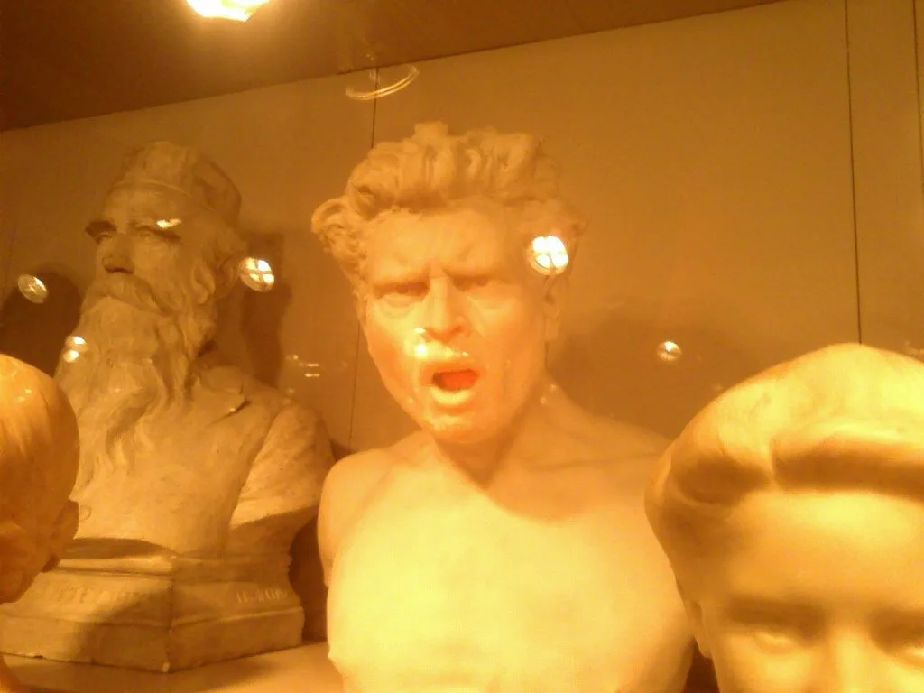 A photo of several marble busts in a museum display case, illuminated by warm lighting that creates reflections on the…