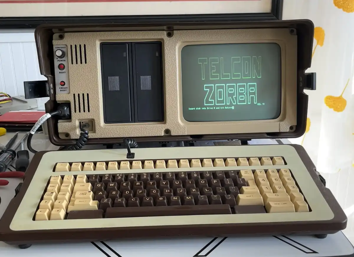 A vintage beige and brown portable computer with a built-in screen displaying the text 'TELCON ZORBA' and the instruction…