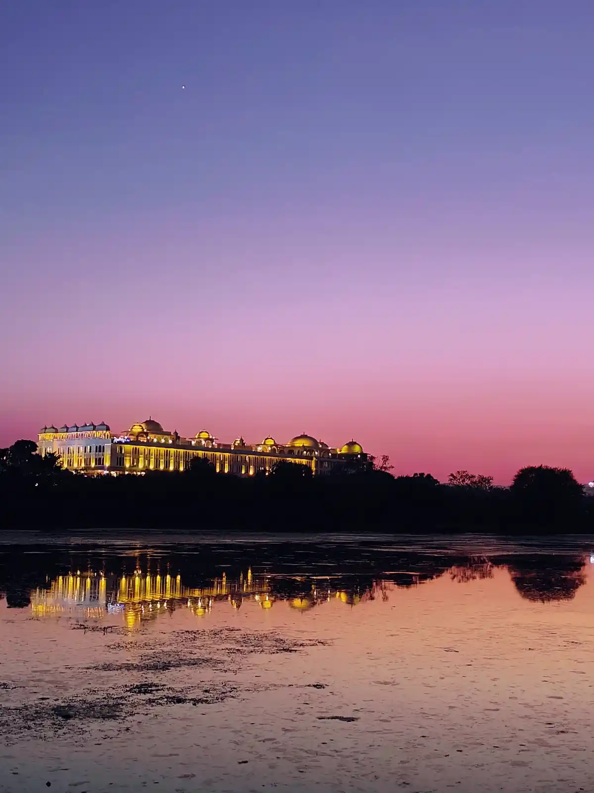A scenic photo of a grand, illuminated palace-style building at dusk, its lights reflecting in a calm body of water below…